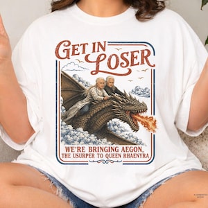 Puede incluir: Una camiseta blanca con un estampado gráfico de dos figuras de pelo largo y rubio montadas sobre un gran dragón que escupe fuego entre nubes. El texto dice: GET IN LOSER WE'RE BRINGING AEGON, THE USURPER TO QUEEN RHAENYRA, en un marco azul.