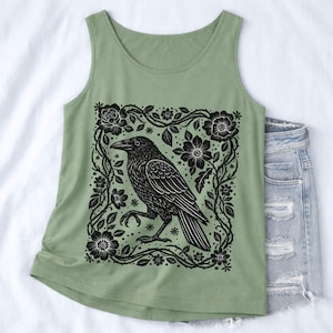 Puede incluir: Una camiseta sin mangas de color verde salvia con un estampado gráfico negro de un cuervo de pie dentro de un marco cuadrado de enredaderas y flores. El diseño está centrado en la parte delantera y muestra líneas detalladas.