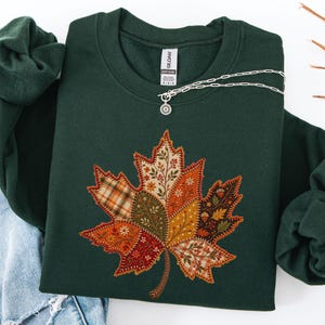 Puede incluir: Una sudadera de cuello redondo en color verde bosque oscuro con un gran gráfico central de hoja de arce compuesto por varios segmentos estilo patchwork. El diseño incluye patrones de cuadros, motivos florales y bellotas en tonos naranja, marrón y crema.