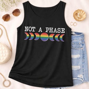 Puede incluir: Una camiseta de tirantes negra con el texto blanco NOT A PHASE sobre un gráfico de las fases lunares en colores arcoíris. El diseño incluye un círculo completo y formas de media luna en una fila horizontal en el centro de la prenda.