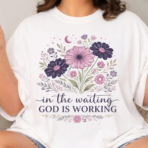 以下が含まれることがあります： 白いTシャツに花の模様。ピンクと紫の花、緑の葉、そして「in the waiting GOD IS WORKING」の文字がデザインされています。三日月と小さな花も含まれています。