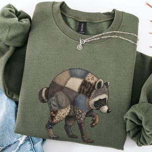 Puede incluir: Una sudadera de cuello redondo color verde oliva con un gráfico de un mapache compuesto por varios parches de tela en tonos marrón, azul y beige. La sudadera tiene cuello y puños acanalados, con un collar de cadena plateada en el escote.