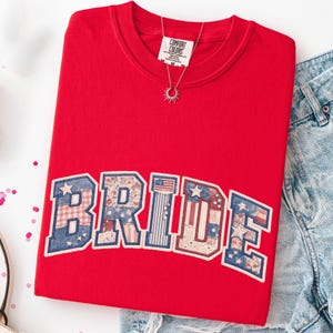 Puede incluir: Una sudadera roja Comfort Colors con la palabra "BRIDE" en un diseño de patchwork con estrellas, rayas y estampados florales. Se ve un collar plateado con un colgante de sol. La sudadera se combina con pantalones cortos de mezclilla.