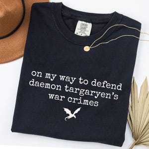 Peut inclure: Un t-shirt noir à manches courtes avec le texte imprimé en blanc "on my way to defend daemon targaryen's war crimes" et un petit graphique représentant la silhouette d'un dragon blanc. L'étiquette Comfort Colors est visible au col.