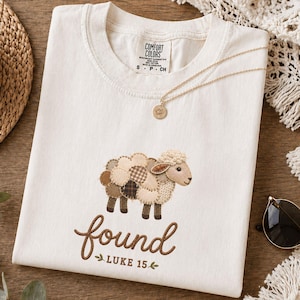 Puede incluir: Una camiseta de manga corta color blanco roto con un gráfico de una oveja hecha de varios parches con patrones marrones y beige. Debajo de la oveja, el texto dice "found" en una fuente cursiva marrón, seguido de "LUKE 15" y dos pequeñas hojas verdes.