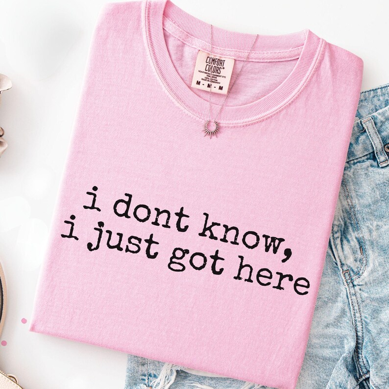 Puede incluir: Una camiseta de color rosa claro con el texto en negro "i dont know, i just got here" impreso en la parte delantera. La prenda tiene cuello redondo y una etiqueta de la marca Comfort Colors en el interior, junto a unos vaqueros azules.