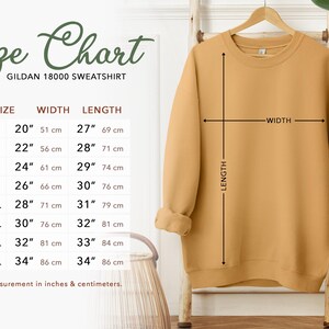Puede incluir: Una sudadera Gildan 18000 de color amarillo mostaza en una percha de madera. A la izquierda hay una tabla de tallas que enumera desde la S hasta la 5XL con las medidas de ancho y largo en cent&iacute;metros, que van de 51 a 86 cm.