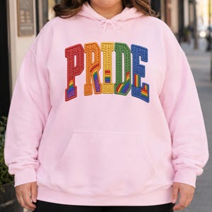 Puede incluir: Una sudadera con capucha de color rosa claro que presenta la palabra PRIDE bordada en letras grandes de estilo bloque en el pecho. Cada letra está rellena con un patrón de arcoíris texturizado y multicolor, e incluye un bolsillo tipo canguro.