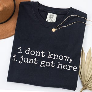 Puede incluir: Una camiseta negra de manga corta con el texto blanco "i dont know, i just got here" impreso en la parte delantera. La camiseta tiene una etiqueta de la marca Comfort Colors en el cuello interior, indicando una talla small, 100% algod&oacute;n.