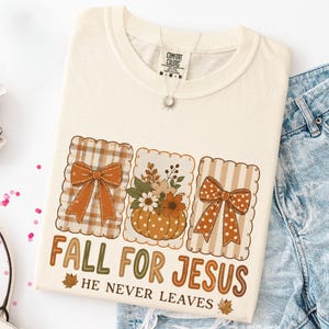 May include: A cream-colored t-shirt featuring a graphic with three scalloped rectangular frames. The outer frames contain orange bows, while the center frame displays a pumpkin with flowers. The text reads FALL FOR JESUS HE NEVER LEAVES.