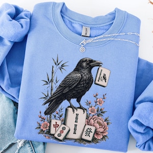 May include: A light blue crewneck sweatshirt featuring a graphic of a black crow perched on a mahjong tile. The crow holds another tile in its beak, surrounded by bamboo stalks, pink cherry blossoms, and a large peony flower illustration.