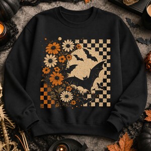 May include: A black crewneck sweatshirt featuring a graphic print of orange and white daisies, black silhouettes of flying bats, and a beige checkered pattern. The design is centered on the chest, combining floral elements with a dark theme.