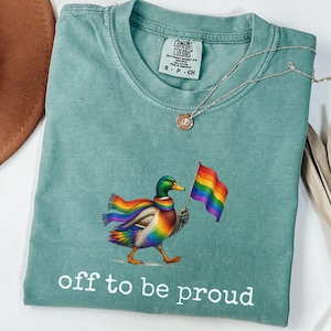 Puede incluir: Una camiseta de manga corta color verde espuma de mar con un gráfico de un pato real que lleva una bufanda arcoíris y sostiene una bandera arcoíris. El texto "off to be proud" está impreso en blanco debajo del pato. Etiqueta Comfort Colors.