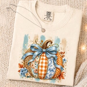 May include: A cream-colored t-shirt featuring a graphic of a pumpkin decorated with leopard print, orange gingham, floral patterns, and polka dots. A large blue ribbon is tied around the pumpkin, surrounded by blue and orange autumn leaves.