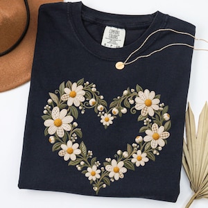 May include: A black short-sleeved t-shirt featuring an embroidered heart-shaped wreath made of white daisies with yellow centres, green leaves, and small buds. The shirt has a Comfort Colors brand label at the neck and a thin gold chain necklace.