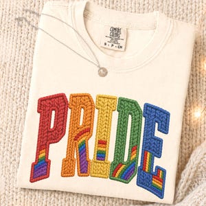 Puede incluir: Una camiseta color crema con la palabra PRIDE bordada en letras grandes y texturizadas con estilo de punto trenzado. Cada letra muestra una franja de acento con los colores del arcoíris. Un collar plateado fino con un dije redondo descansa sobre ella.
