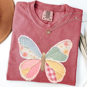 May include: A dusty rose Comfort Colors t-shirt with a patchwork butterfly design. The butterfly wings feature various pastel patterns, including floral and gingham. A silver necklace with a small pendant rests on the shirt.