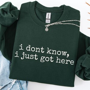 Puede incluir: Una sudadera de cuello redondo en color verde bosque oscuro con el texto blanco "i dont know, i just got here" impreso en una fuente estilo m&aacute;quina de escribir. La prenda incluye una etiqueta de la marca Gildan y un collar plateado.