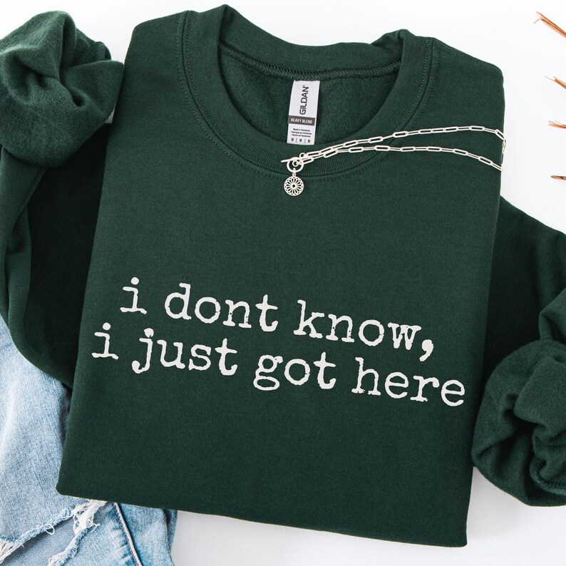 Puede incluir: Una sudadera de cuello redondo en color verde bosque oscuro con el texto blanco "i dont know, i just got here" impreso en una fuente estilo m&aacute;quina de escribir. La prenda incluye una etiqueta de la marca Gildan y un collar plateado.