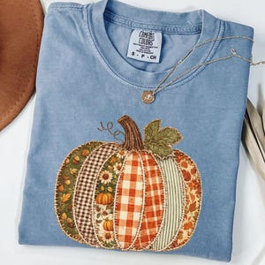 May include: A blue short-sleeve t-shirt with a Comfort Colors label at the neck. The front features a large pumpkin graphic composed of various fabric-style patches in plaid, floral, and leaf patterns. A thin gold chain necklace rests on the shirt.
