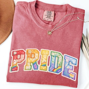May include: A dusty rose short-sleeve t-shirt featuring the word PRIDE in large, quilted-style block letters. Each letter displays a unique pattern including floral prints, gingham, and rainbows. A thin silver chain necklace rests on the neckline.