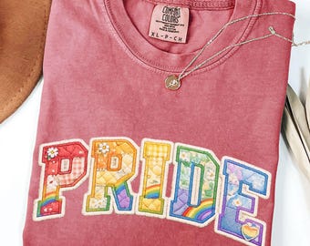 May include: A dusty rose-colored t-shirt with the word "PRIDE" appliqued across the front in a patchwork design. The letters are filled with floral and rainbow patterns. A silver necklace with a small pendant rests on the shirt.