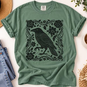 Puede incluir: Una camiseta de manga corta color verde salvia con un estampado gráfico negro de un cuervo de pie dentro de un marco cuadrado de intrincadas enredaderas florales y flores. La camiseta tiene cuello redondo y etiqueta Comfort Colors.