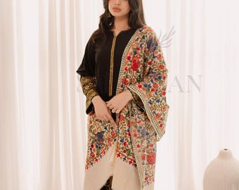 May include: A woman wearing a black outfit with a colorful floral shawl. The shawl features a cream-colored base with intricate embroidery in red, blue, and green. The outfit includes black pants and a long-sleeved top. The background has white curtains and decorative vases.
