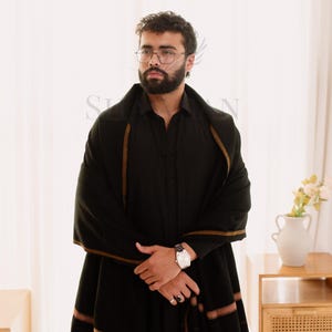May include: A black shawl with brown stripes and fringe is draped over a black shirt. The shawl is worn by a person wearing glasses and a watch. The background is a white curtain and a wooden table.