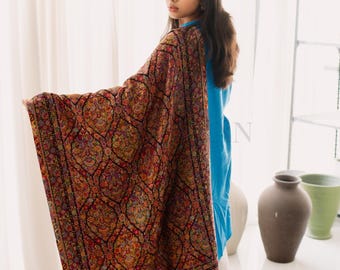 May include: A woman wearing a shawl with an intricate, colorful floral pattern. The shawl is primarily red, with accents of yellow, orange, and green. The woman is wearing a blue top and white pants.