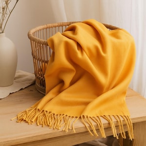 May include: A mustard yellow throw blanket with fringe detail draped over a wooden table. The blanket is soft and textured, with a woven basket in the background. The blanket is approximately 60 inches long.