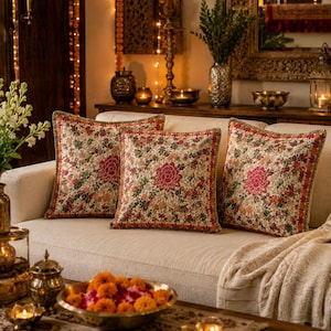 May include: Three square cushions with floral patterns in red, green, and orange on a cream sofa. The cushions have a gold trim and a large floral design in the centre. The room has warm lighting and decorative accents.