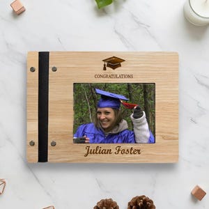 May include: A wooden photo frame with a graduation theme. The frame has a black spine and holds a photo of a person in a graduation gown and cap. The frame is engraved with "Congratulations" and "Julian Foster".