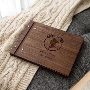 May include: A wooden photo album with a graduation cap design and the text "Lucas Gray Class of 2026". The album is brown and rests on a gray surface with a cream-colored knitted blanket.