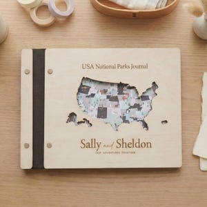 以下が含まれることがあります： 「USA National Parks Journal」の文字と、アメリカ合衆国の地図の切り抜きが入った木製ジャーナル。「Sally and Sheldon」の名前と「OUR ADVENTURES TOGETHER」のフレーズが含まれています。