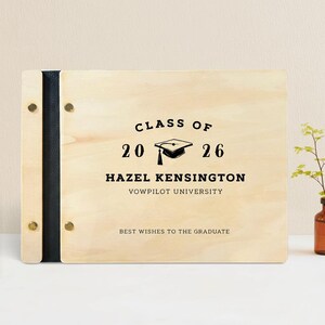 May include: A wooden graduation memory book with black text that reads "Class of 2026" and the graduate's name, Hazel Kensington, and university. The book has a black spine and is held together with gold-colored rivets.