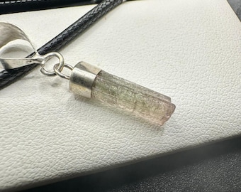 May include: A pendant necklace with a natural, elongated gemstone, silver-coloured metal cap and bail. The gemstone has green and pink hues. The necklace has a black, braided leather cord.