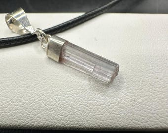 May include: A pendant necklace with a clear, cylindrical gemstone, silver-coloured metal cap and bail. The gemstone has a subtle pink hue. The pendant hangs from a black cord.