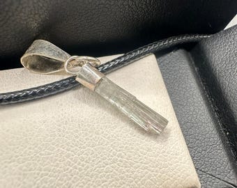 May include: A clear, light green crystal pendant with a silver bail, on a black leather cord. The crystal is rectangular and faceted. The pendant rests on a white textured surface, inside a black box.
