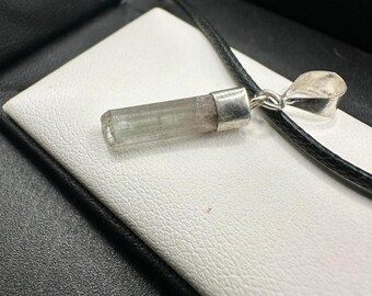 May include: A pendant necklace with a clear, cylindrical gemstone, silver cap and bail. The gemstone has a pale green hue with a hint of pink. The pendant hangs from a black cord. The gemstone is approximately 2 cm long.