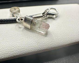 May include: A clear, pink and green tourmaline crystal pendant with a silver bail and clasp. The crystal is mounted on a black cord. The pendant is a natural gemstone, suitable for jewelry.