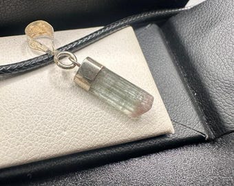 May include: A silver pendant featuring a natural, elongated, light green and pink tourmaline gemstone. The pendant hangs from a black cord, displayed on a white surface inside a black case. The gemstone is polished and cylindrical.