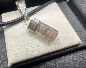 May include: A pendant necklace with a tourmaline gemstone in pink and green. The rectangular gemstone is set in silver-coloured metal. The necklace has a black cord.