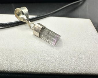 May include: A silver pendant featuring a clear and purple rectangular gemstone. The pendant hangs from a black cord. The gemstone is set in silver, with a loop at the top for the cord to pass through.