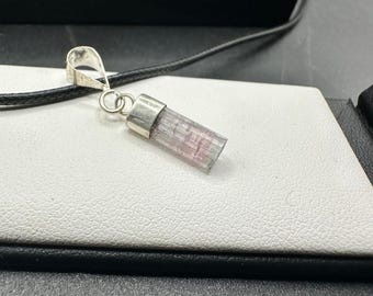May include: A silver pendant with a natural, pink and clear tourmaline crystal. The crystal is rectangular and has a silver cap and loop for the necklace. The pendant hangs on a black cord.