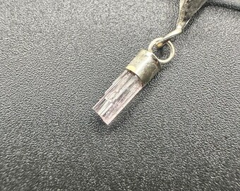 May include: A pendant necklace with a light pink, crystal gemstone. The gemstone is set in silver-coloured metal and hangs from a black cord. The pendant has a simple, elegant design.