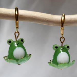 May include: A pair of green frog earrings with gold-colored accents. Each earring features a smiling frog with a white belly, sitting on a lily pad. The earrings are suspended from gold-colored leverback closures.