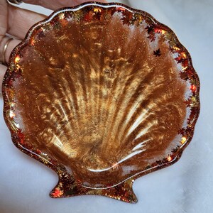 May include: A seashell-shaped resin dish with a shimmering gold interior. The rim is decorated with small, colorful autumn leaves. The dish is a decorative item, perfect for holding small items.