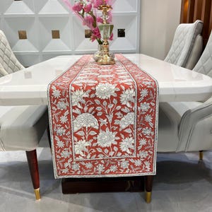 May include: A red and white floral table runner is centered on a white marble dining table. The runner features a repeating pattern of large white flowers and leaves on a red background. The table is surrounded by white upholstered chairs.