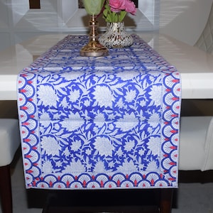 Può includere: Runner da tavolo blu e bianco a fantasia floreale con bordo smerlato con accenti rossi. Il runner è posto su un tavolo bianco, con sedie bianche su entrambi i lati. Un candelabro dorato e un vaso con fiori sono sul tavolo.